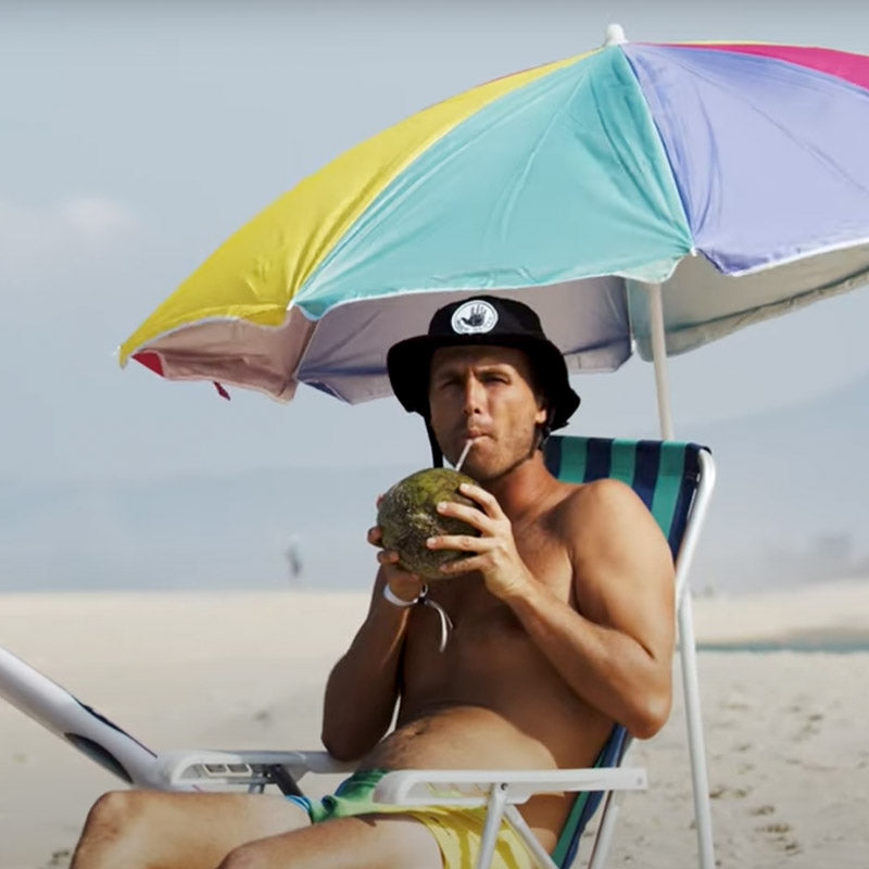 Jesse Mendes sipping from a coconut on a beach with a colorful umbrella