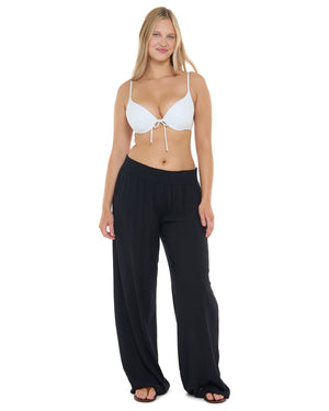 A woman modeling a two-piece swimwear set consisting of a white triangle bikini top with a front tie and black wide-leg swim pants, shot against a solid white background.